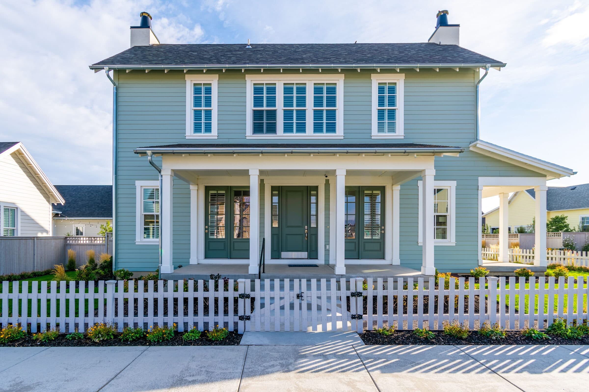 White picket fence with a double walk gate in front of a sage-green craftsman home. Material not confirmed.